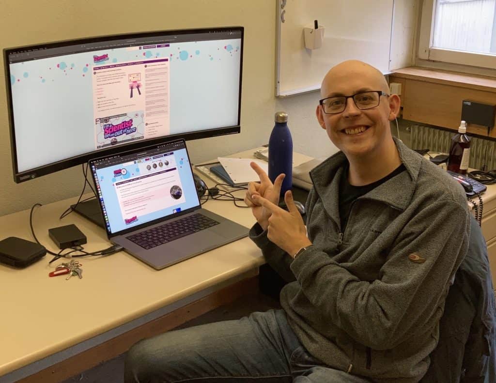 An image of a man sitting down, smiling and pointing at a computer screen.